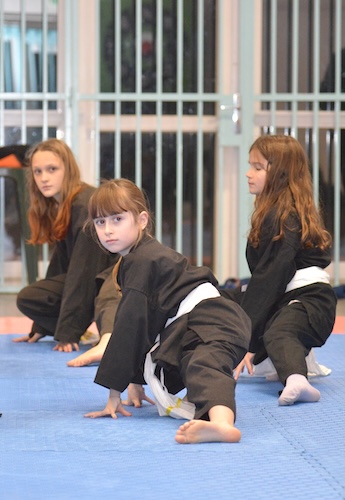 Trois enfants d'environ 8 ans en kimono noir, en train d'exécuter une posture de coup de pied au ras du sol.